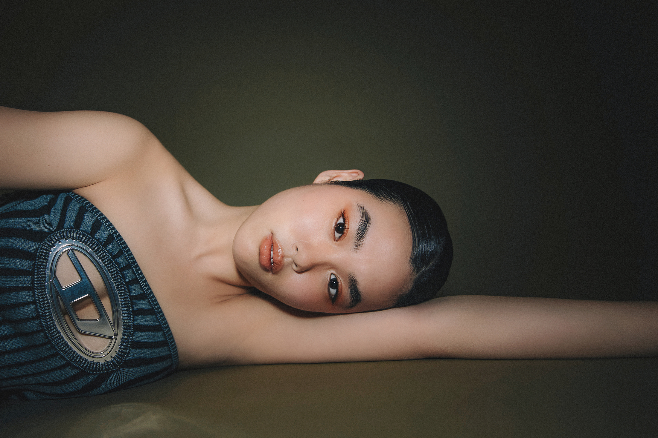 A model laying on an olive green background wearing a slim-fit blended cotton bouclé bandeau top plated with Lycra. A stonewashed finish imparts a worn-in look, whilst an enamelled metal 'D' plaque on the chest creates a peek-a-boo effect and symbolises Diesel branding.